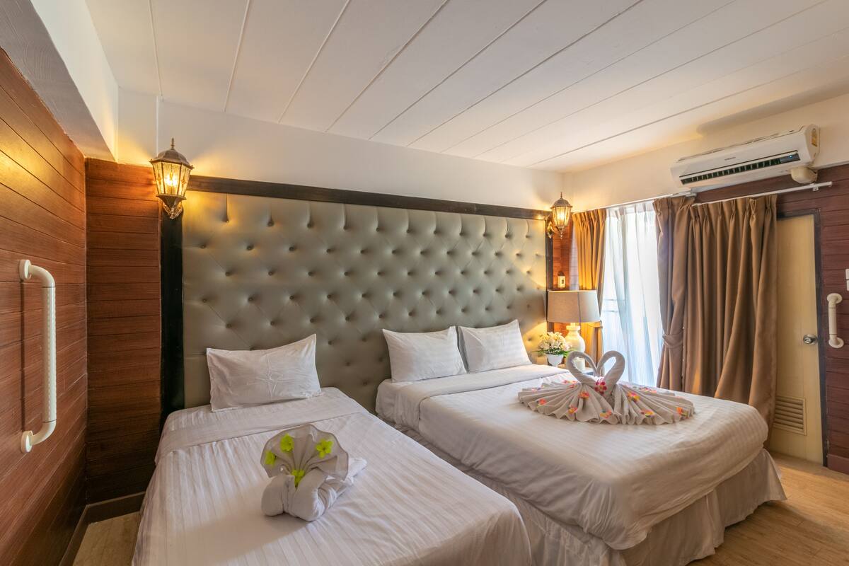 Une chambre d’hôtel avec deux lits, une tête de lit capitonnée, des lampes fixées au mur, un climatiseur et une fenêtre avec des rideaux.