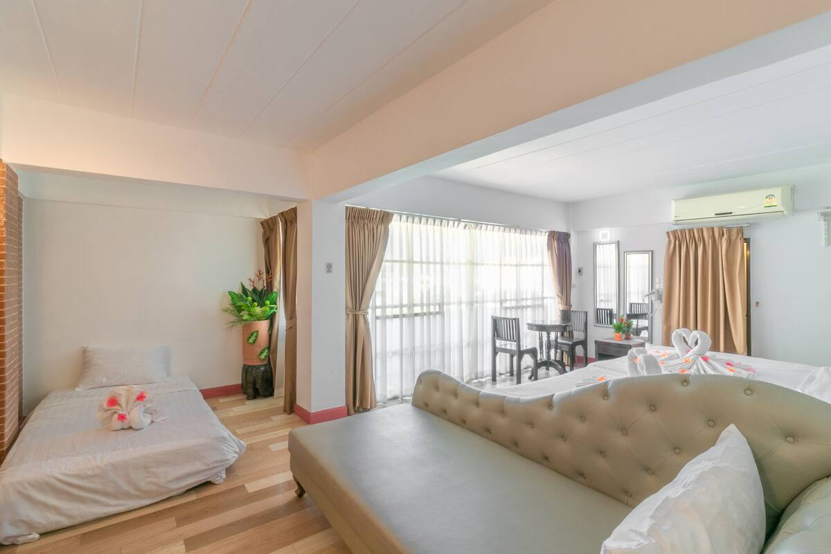 Une chambre d’hôtel avec un grand lit, un lit plus petit, un canapé, un bureau et une chaise. On y trouve également une plante et une fenêtre avec des rideaux.