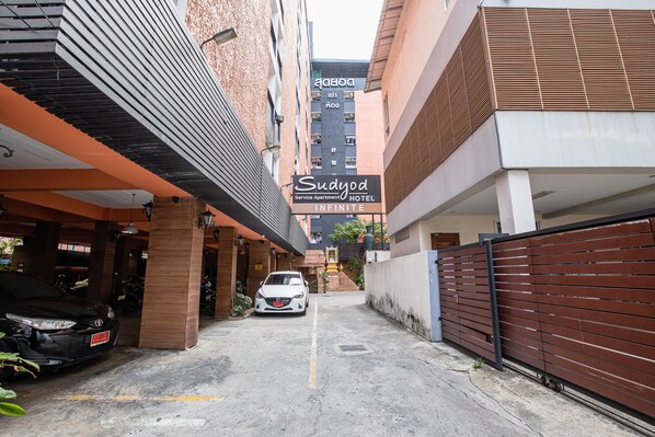 A parking area with a white car and a motorcycle, a sign for 'Sudyod Service Apartment Hotel Infinite', and a building with a brick facade and wooden slats.