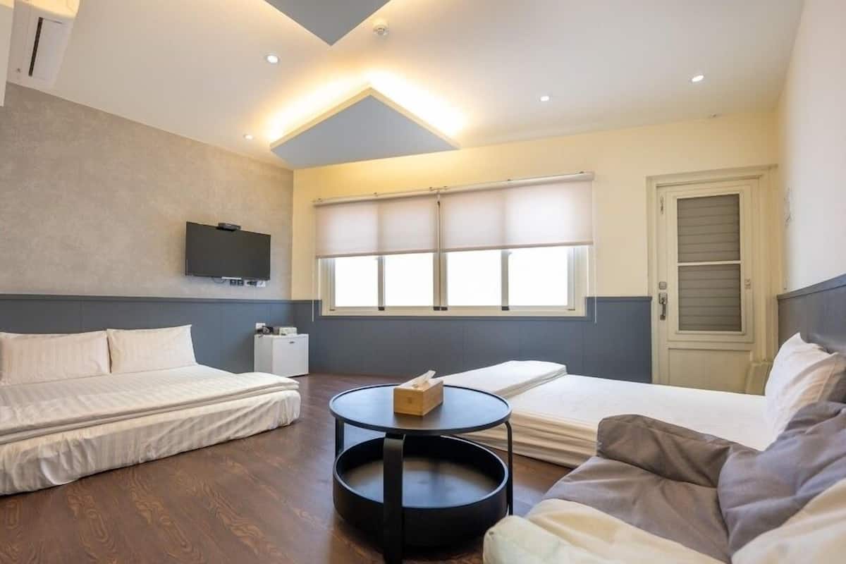 Una habitación de hotel compacta con dos camas, un sofá, una pequeña mesa redonda, un televisor montado en la pared y una ventana con persianas.
