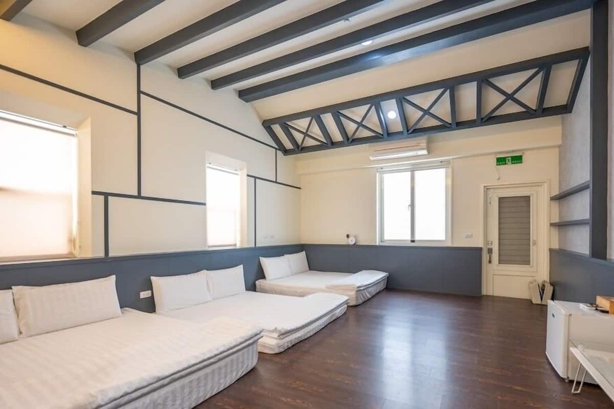 Habitación de hotel con techo alto, vigas a la vista y un ventanal grande. Hay tres camas dispuestas en fila, un escritorio pequeño y una silla.