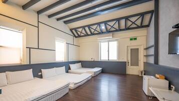 A hotel room with a high ceiling, exposed beams, and a large window. There are three beds arranged in a row, a small desk, and a chair.