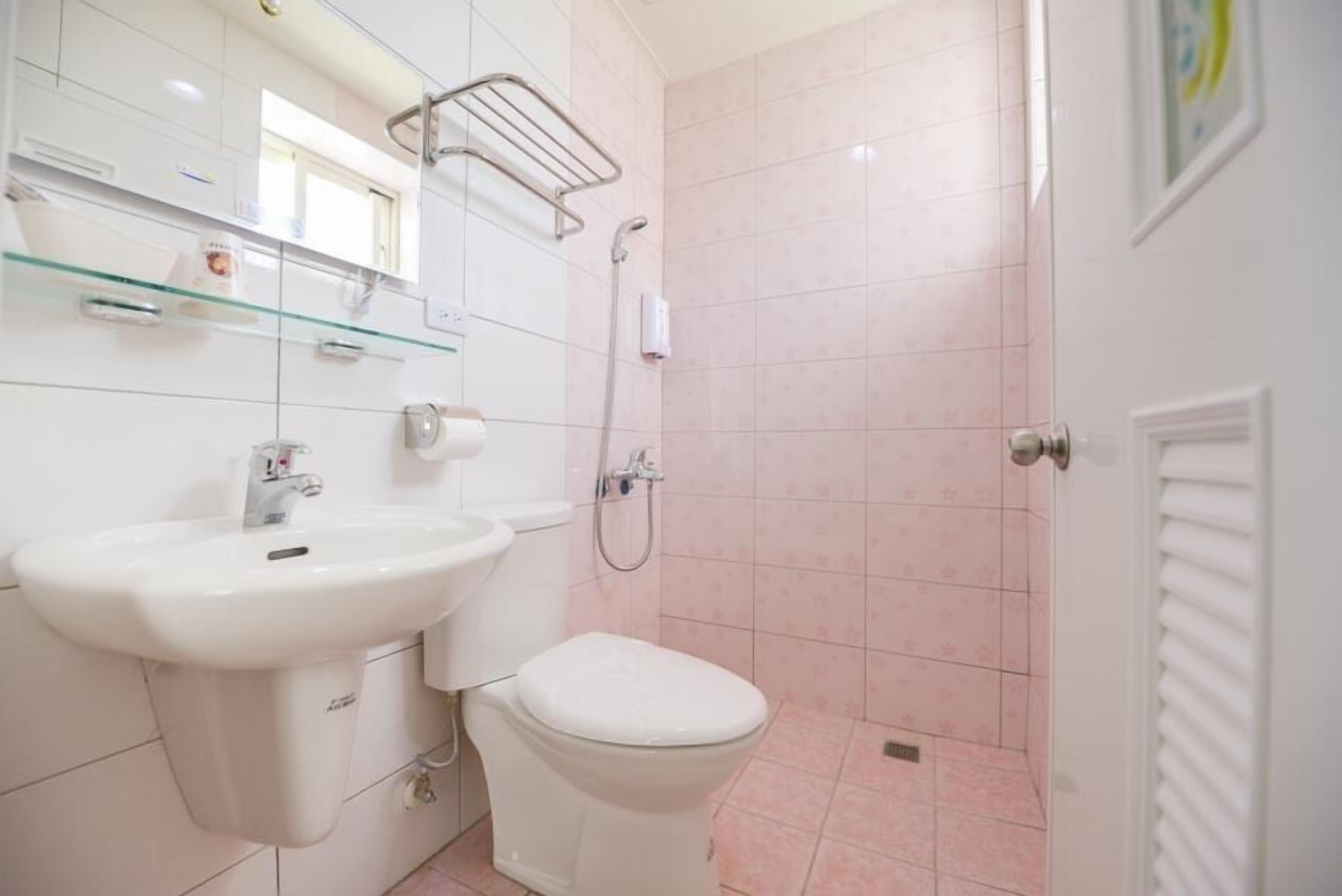 Un baño con lavamanos, inodoro y ducha blancos. Hay una repisa de vidrio sobre el lavamanos y un perchero en la pared.