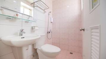 Un baño con lavamanos, inodoro y ducha blancos. Hay una repisa de vidrio sobre el lavamanos y un perchero en la pared.
