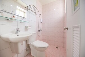 Un baño con lavamanos, inodoro y ducha blancos. Hay una repisa de vidrio sobre el lavamanos y un perchero en la pared.