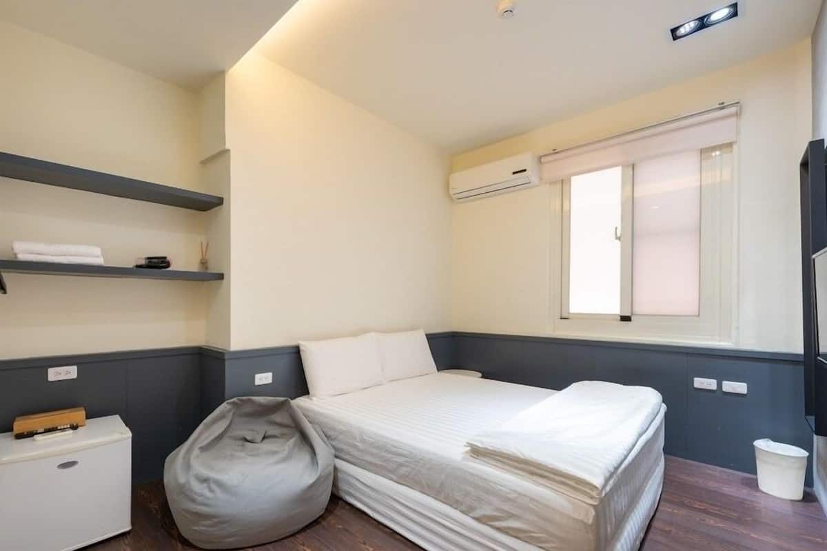 Un dormitorio pequeño y ordenado con cama, un sillón de mimbre, un mueble blanco pequeño y una ventana con persianas.