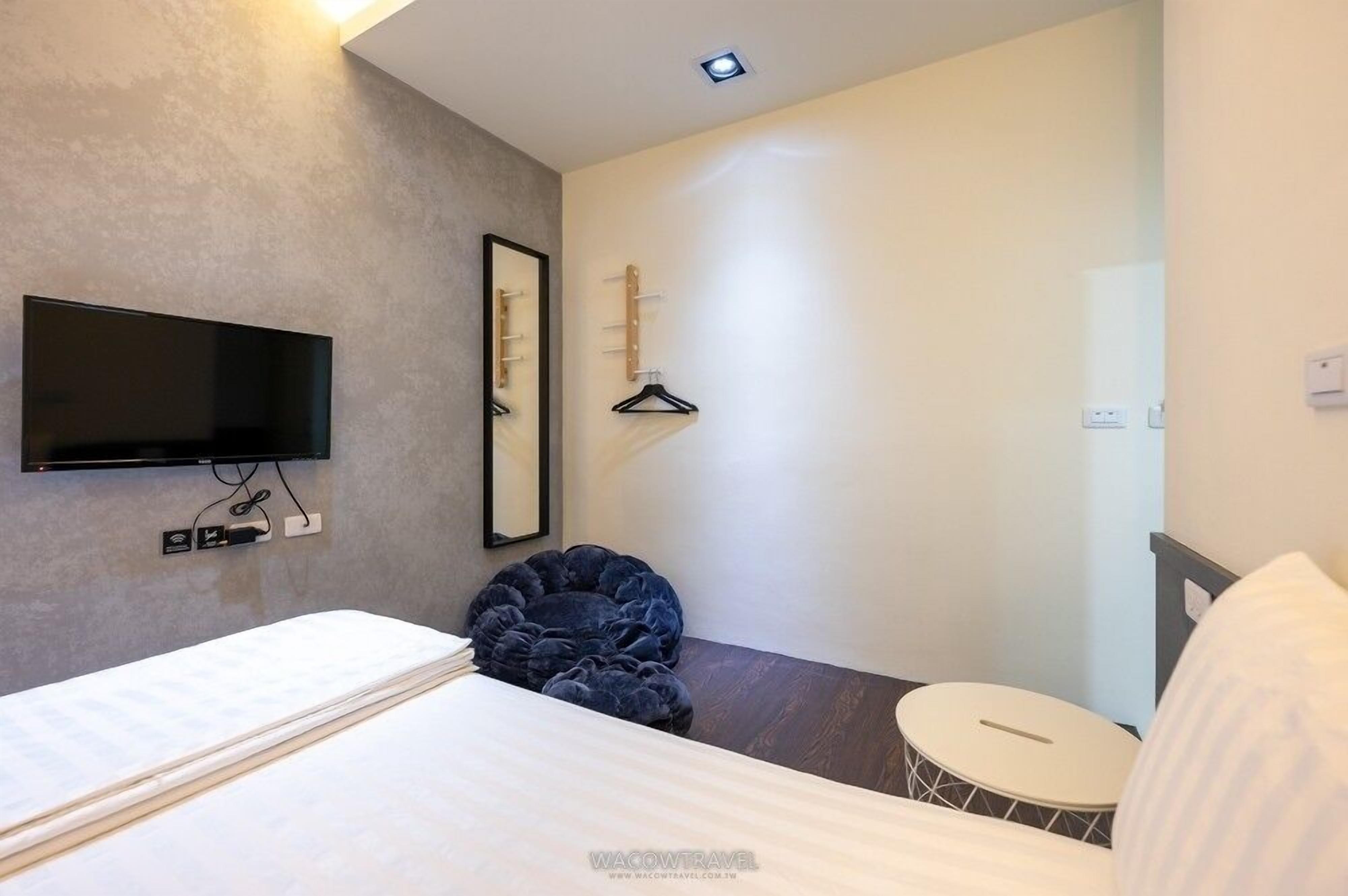 A modern hotel room with a bed, a flat-screen TV mounted on the wall, a mirror, a hanging light fixture, and a small round table.