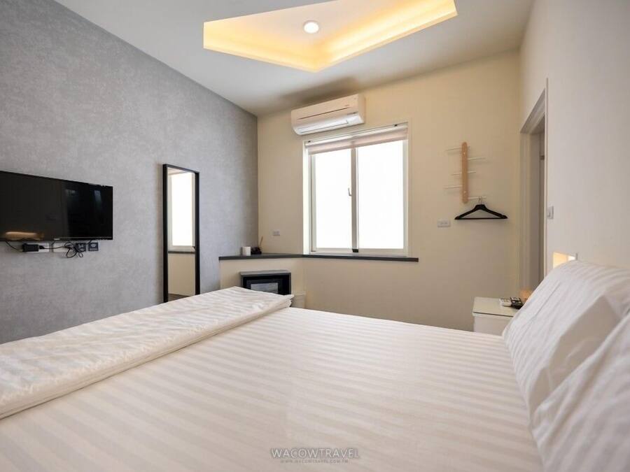 Quarto de hotel moderno com cama, televisão de tela plana na parede, janela com persianas e mesa de cabeceira pequena.