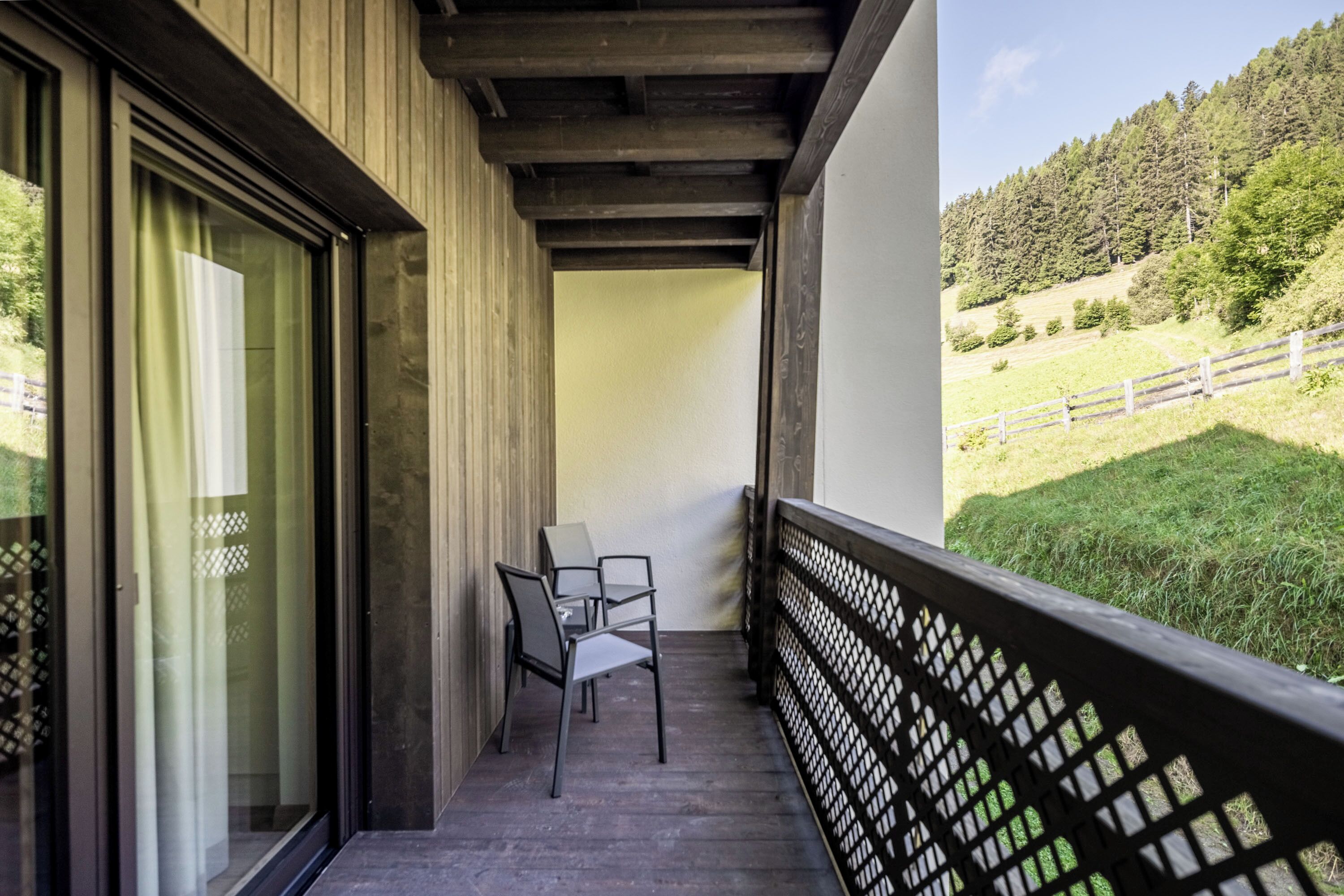 A balcony with a wooden floor, two chairs, and a view of a green landscape with trees and a fence.