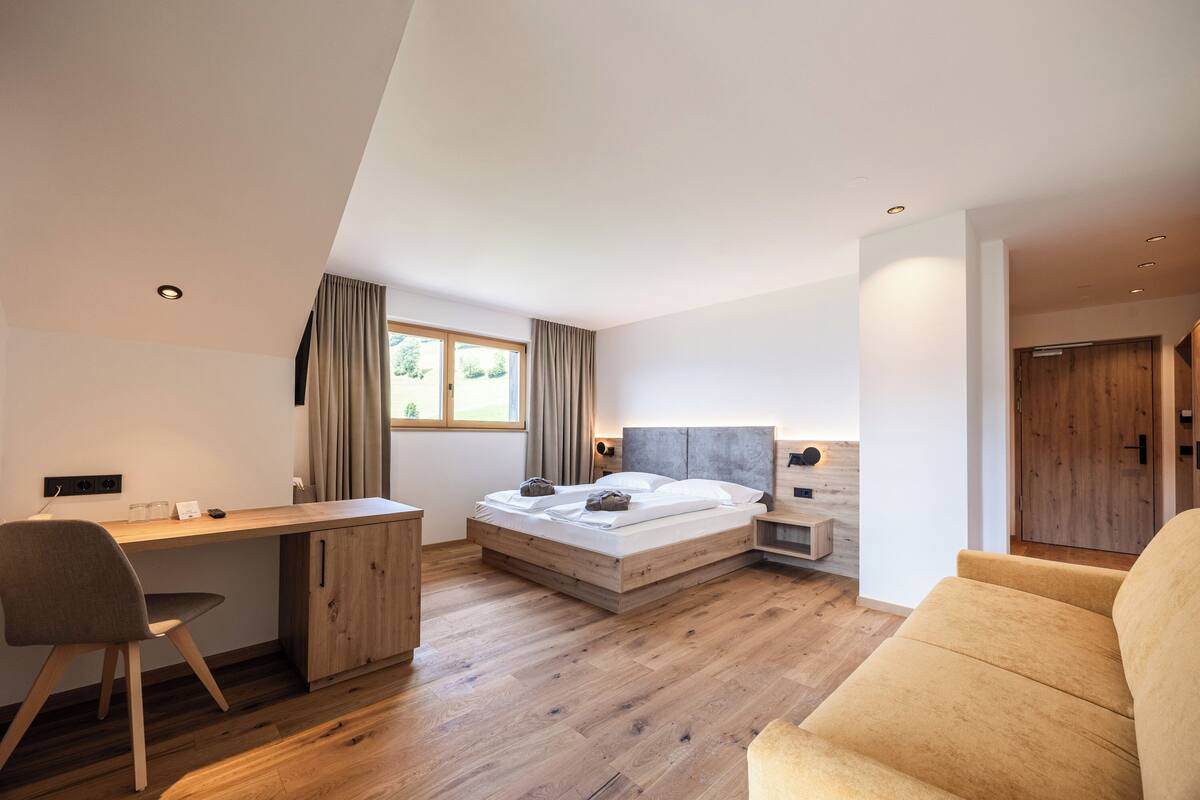 A modern bedroom with a wooden desk, a bed with a headboard, a sofa, and a window with curtains.