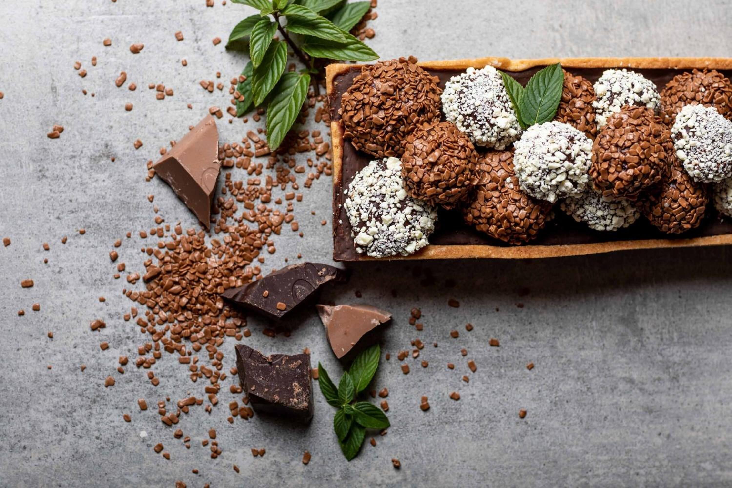 Una bandeja de trufas de chocolate con hojas de menta y trozos de chocolate esparcidos sobre una superficie texturizada.