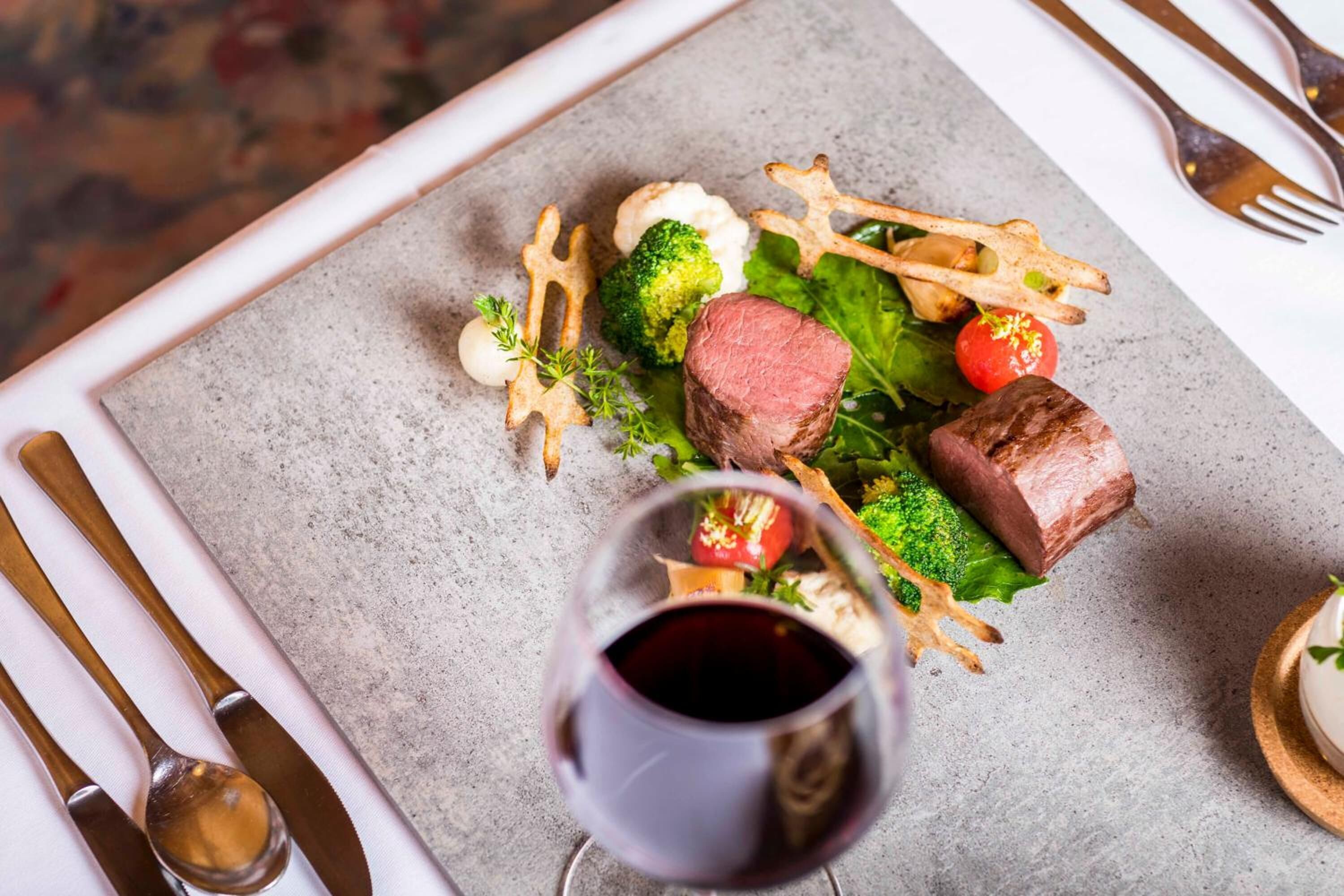 Un plato con carne, brócoli y otras verduras, acompañado de una copa de vino tinto, sobre una mesa con cubiertos.