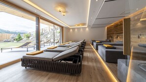 A modern spa room with a long bench seating area, a large window with a view of the outdoors, and a wooden wall with built-in lighting. - Hotel Rommisa (Perca)