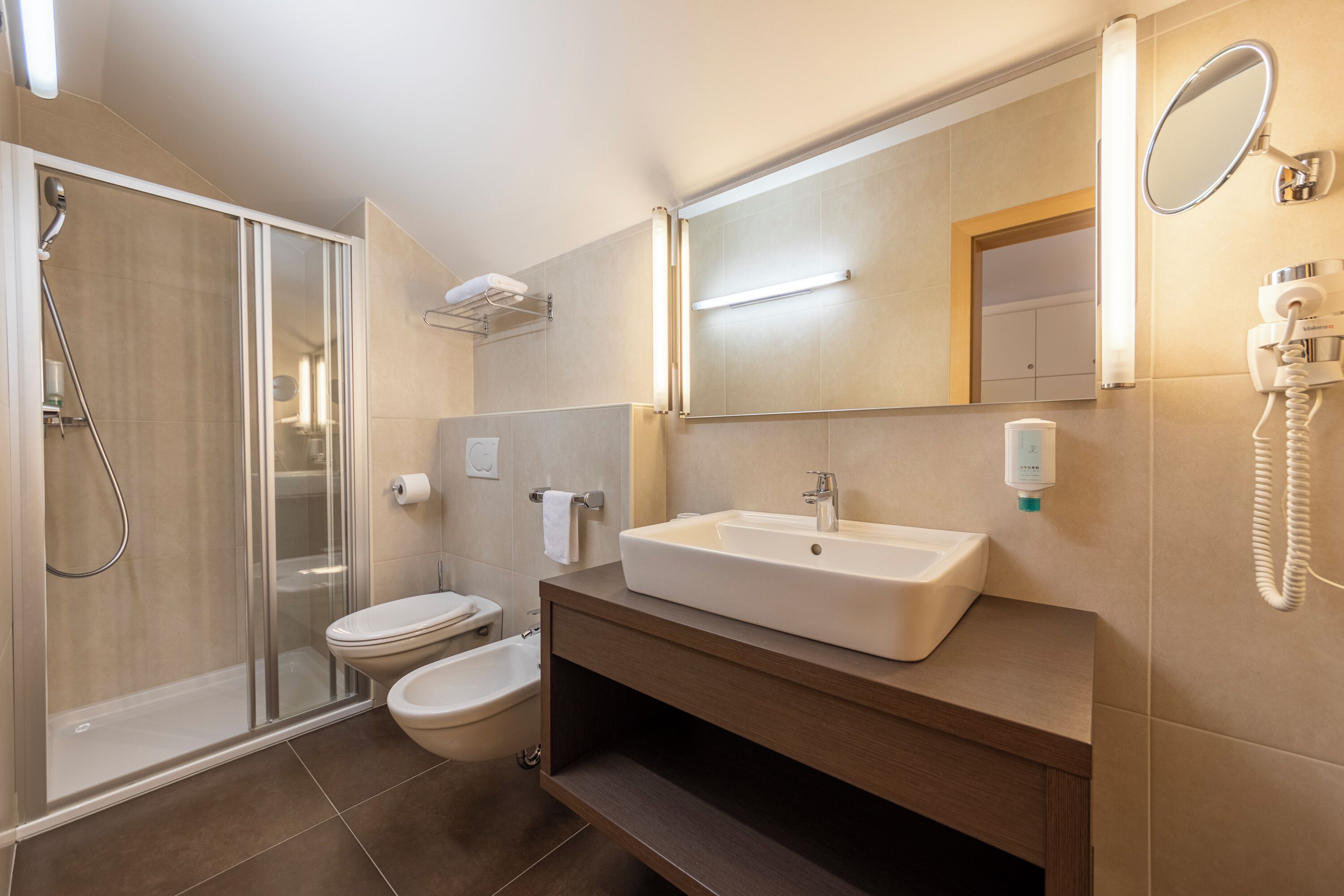 Un baño moderno con ducha envuelta en vidrio, inodoro, bidé, lavamanos con espejo y secador de pelo en la pared.
