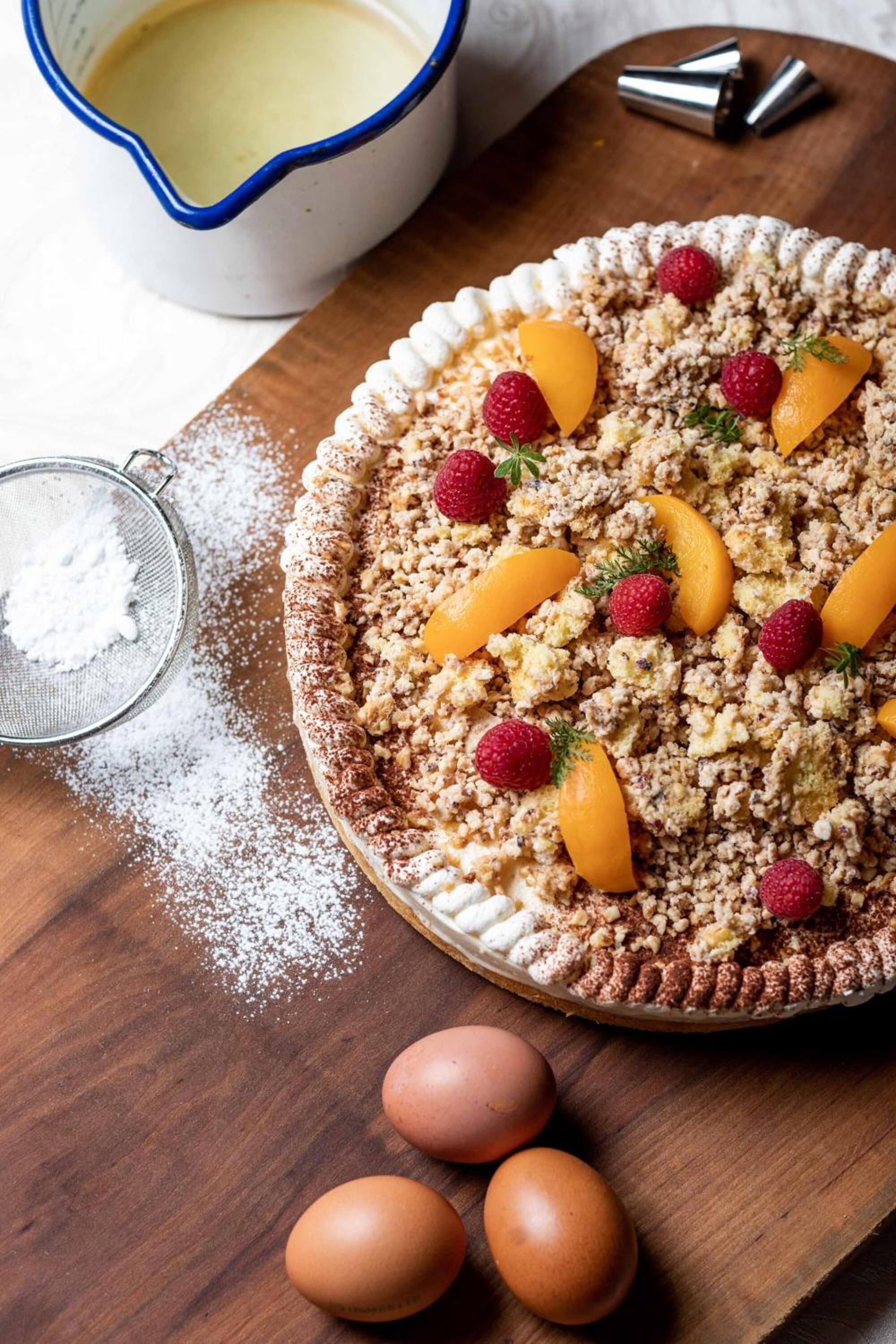A dessert tart on a wooden board, with a bowl of cream and a sieve of powdered sugar nearby.