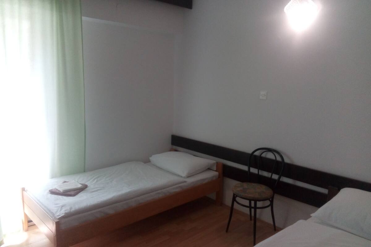 Ein kleines Zimmer mit einem Einzelbett, Holzboden, einem Sessel und einem Nachttisch.