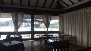 A restaurant with a high ceiling, wooden beams, and large windows with curtains. Tables and chairs are arranged for dining.