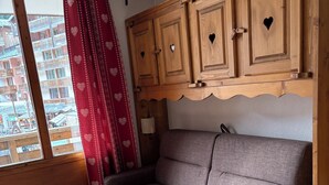 A cozy room with a grey sofa, wooden cabinets with heart cutouts, a window with a red heart-patterned curtain, and a view of buildings outside.
