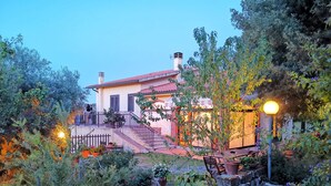 A house with a red roof, a staircase leading to a porch, and a garden with various plants and a bench. - Agriturismo Norianino (Manciano)