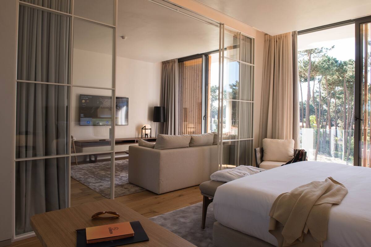 Ein modernes Hotelzimmer mit einem großen Bett, einer Couch, einem Sessel, einem Fernseher und einer Glasschiebetür, die auf einen Balkon mit Blick auf Bäume führt.