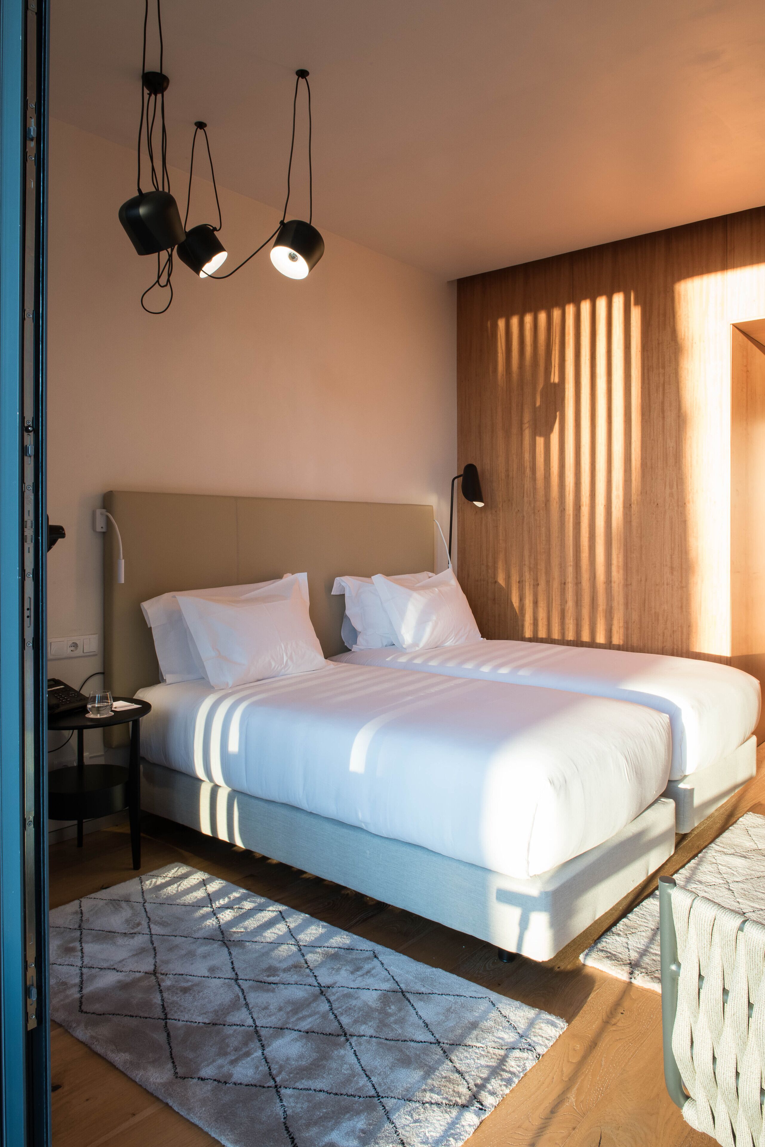 A modern hotel room with a large bed, a bedside table with a lamp, a wooden headboard, and a view of the outside through a window.