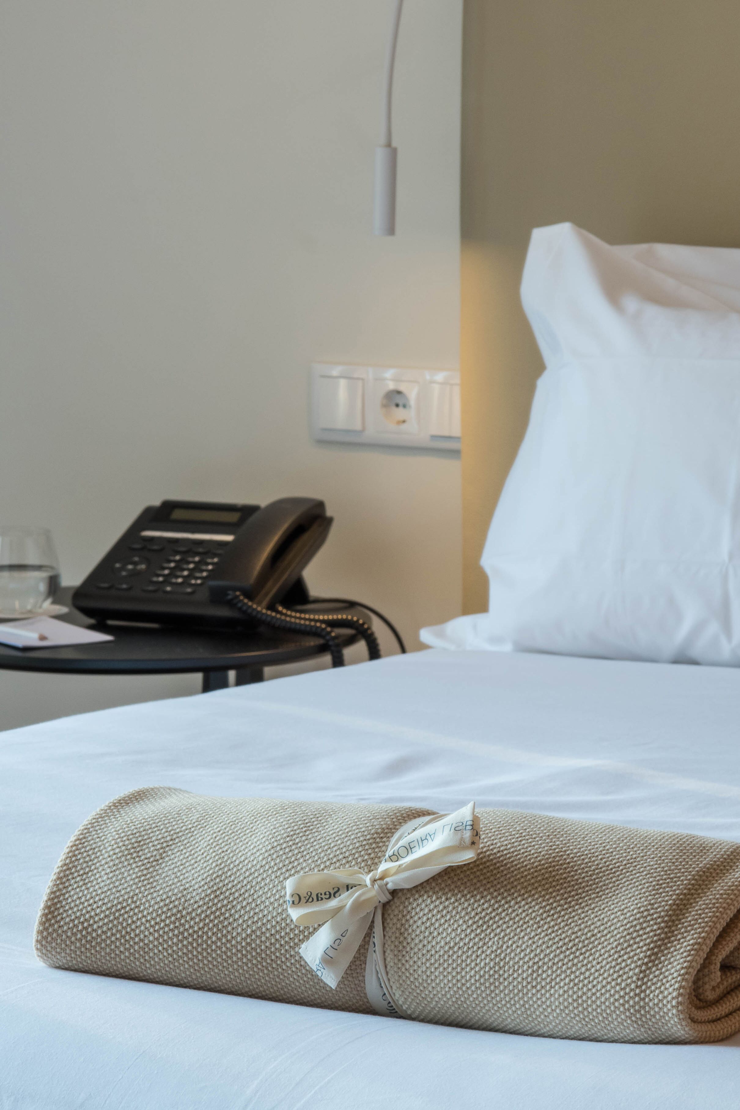 Ein ordentlich bezogenes Bett mit einem zusammengerollten Handtuch und einem schwarzen Telefon auf einem Nachttisch.