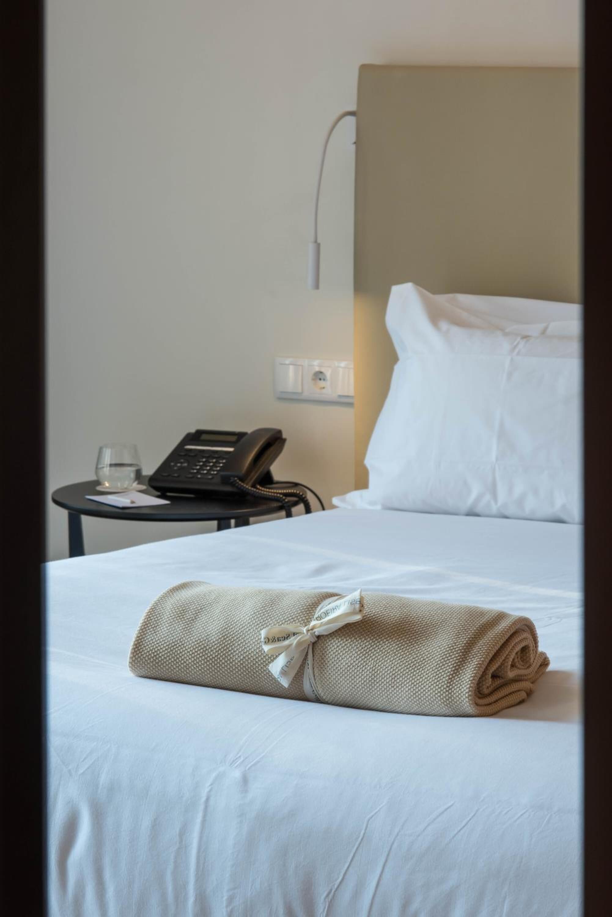 A neatly made bed with a rolled towel and a bedside phone on a small table beside it.