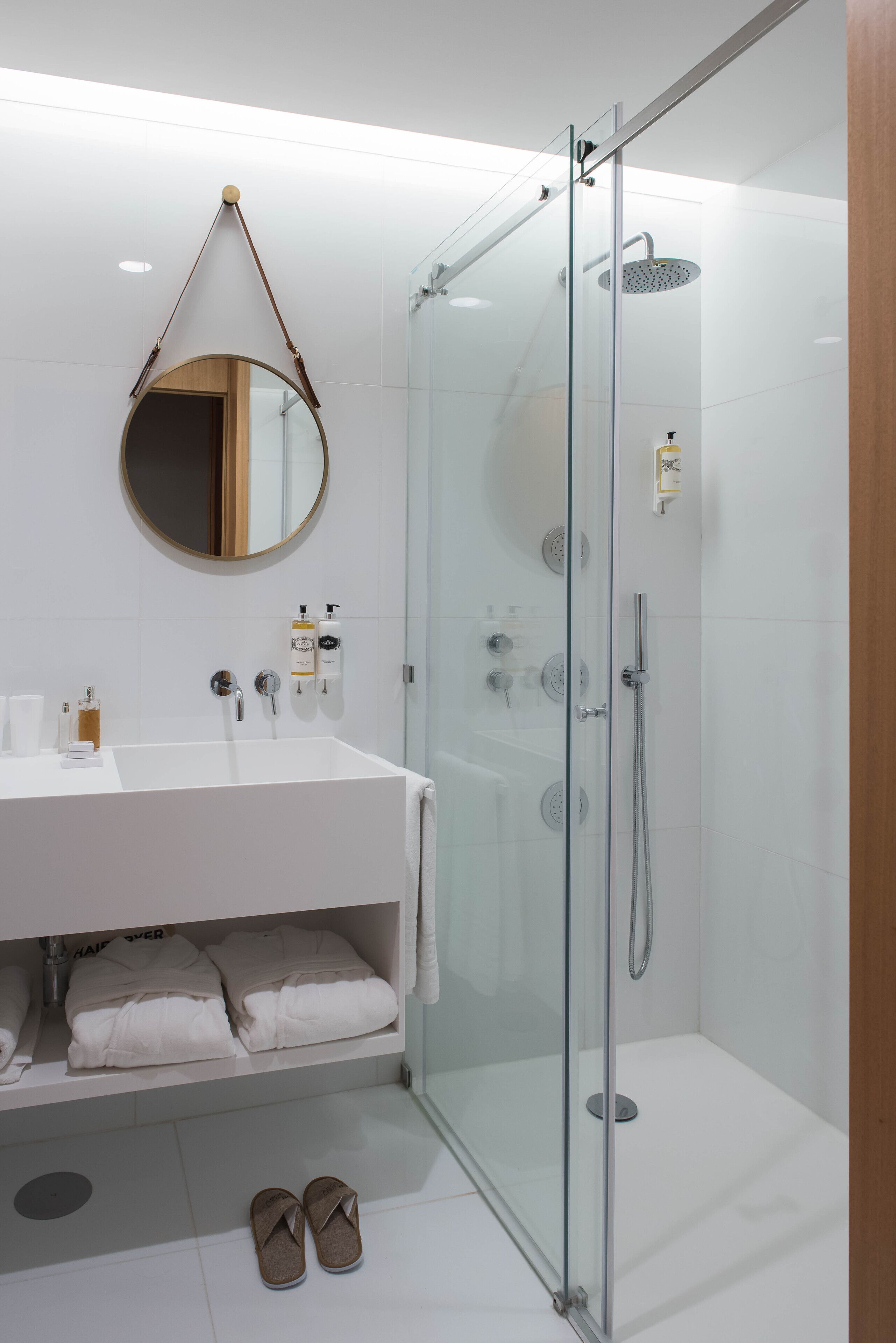 Un baño moderno con ducha envuelta en vidrio, un lavamanos blanco con artículos de aseo y un espejo redondo.