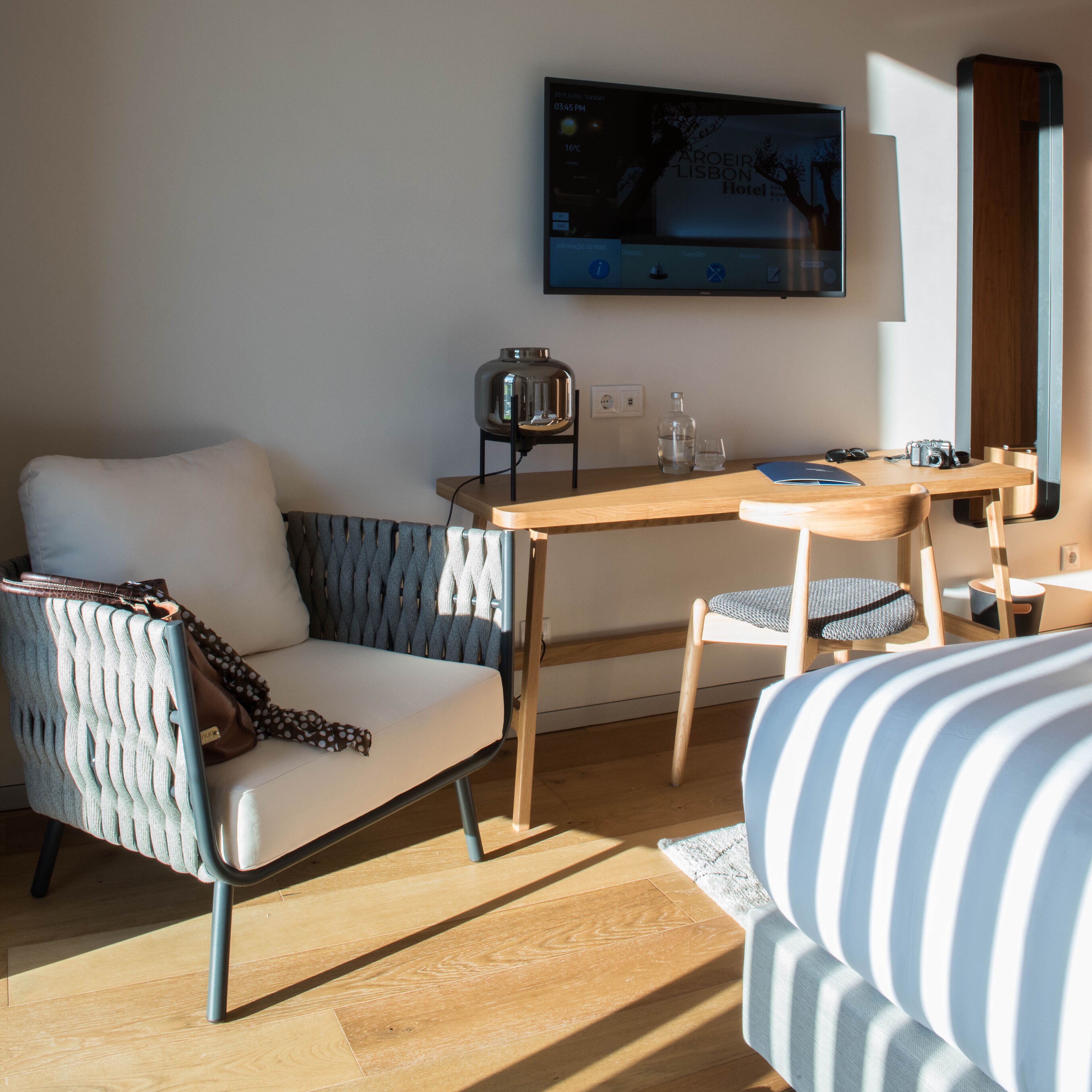 A modern hotel room with a wooden desk, a comfortable sofa, a television mounted on the wall, and a window providing natural light.