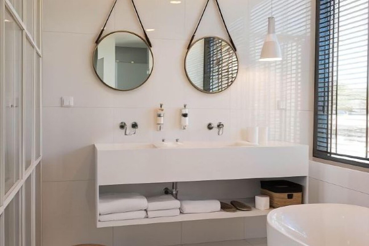 A modern bathroom with a white vanity, two round mirrors, a large bathtub, and a window with blinds.