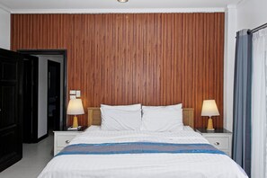 Habitación de hotel con pared de paneles de madera, cama con ropa de cama blanca, dos lámparas de noche y una mesita de noche con control remoto.