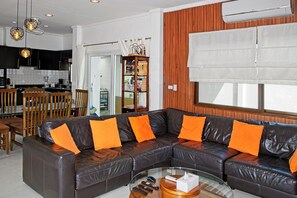 A modern living room with a leather sectional sofa, a glass coffee table, and a kitchen area with black cabinets and a white backsplash.