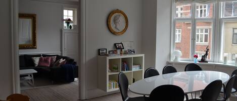 Une pièce avec une table à manger blanche, des chaises noires, une bibliothèque blanche, un tableau encadré et une fenêtre donnant sur des immeubles.