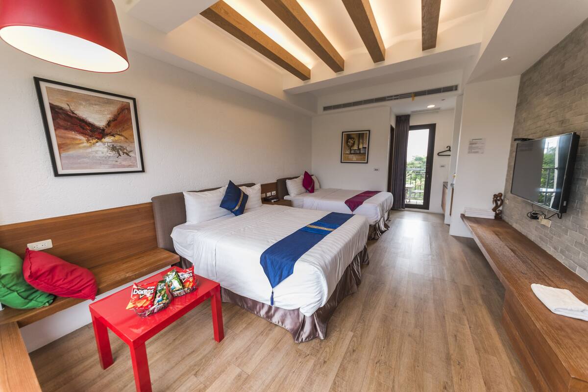 A modern hotel room with two beds, a red table with snacks, a wooden bench, and a TV on the wall.