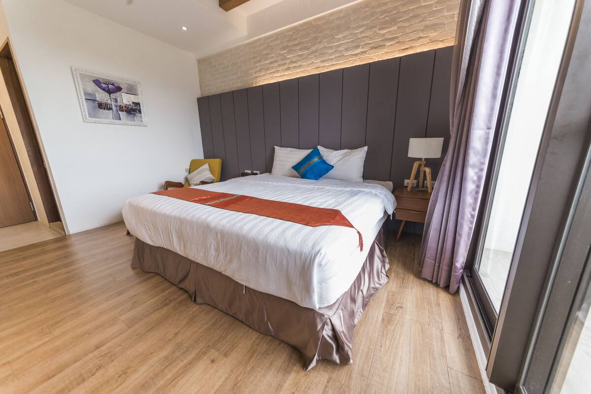 A modern hotel room with a large bed, wooden flooring, a brick accent wall, and a window with curtains.