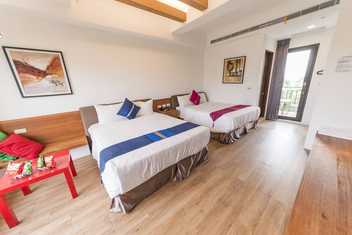 A hotel room with two beds, a wooden headboard, a red table with snacks, and a balcony door.