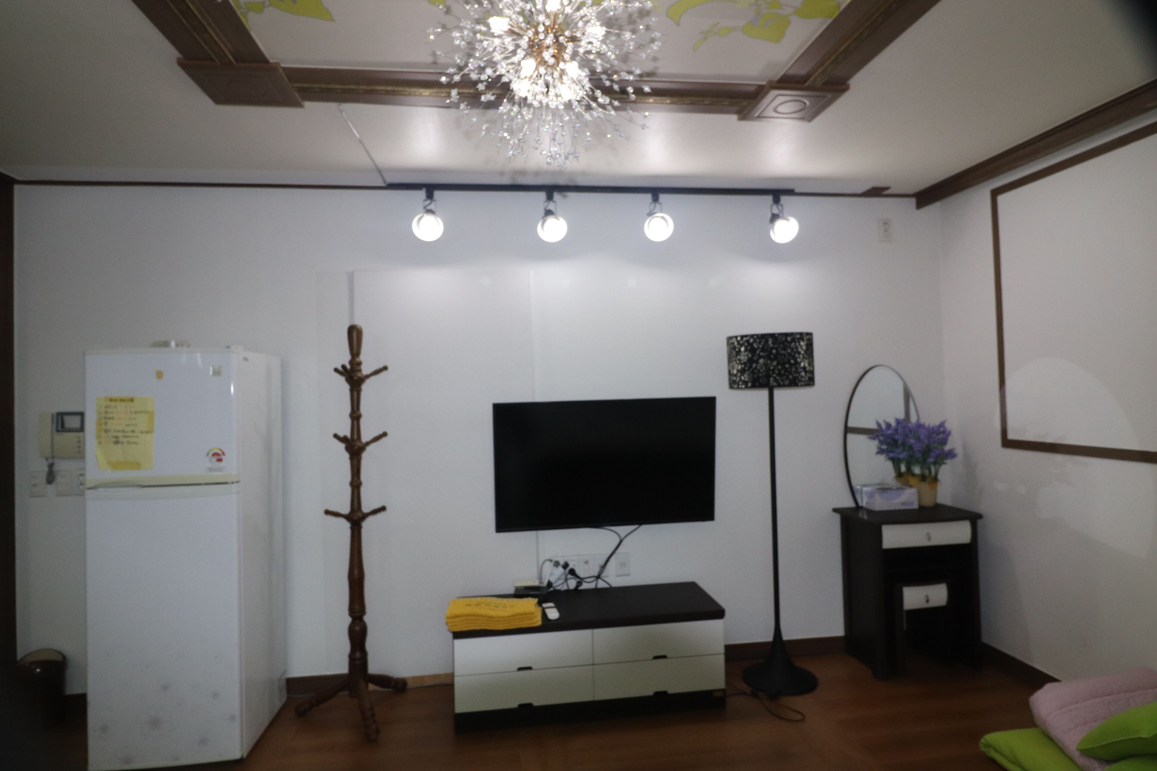 A room with a flat-screen TV mounted on the wall, a white refrigerator, a wooden coat rack, a black floor lamp, and a small desk with a potted plant.