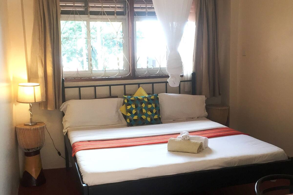 A single bed with a white mattress, a red and white striped bedspread, and a white towel folded at the foot of the bed. There is a bedside lamp on a wooden base, a window with metal bars, and a curtain on the left side.