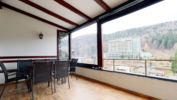 A balcony with wicker chairs and a table, wooden flooring, and a view of a hotel and forested hills. - Apartment Sofia (Sinaia)