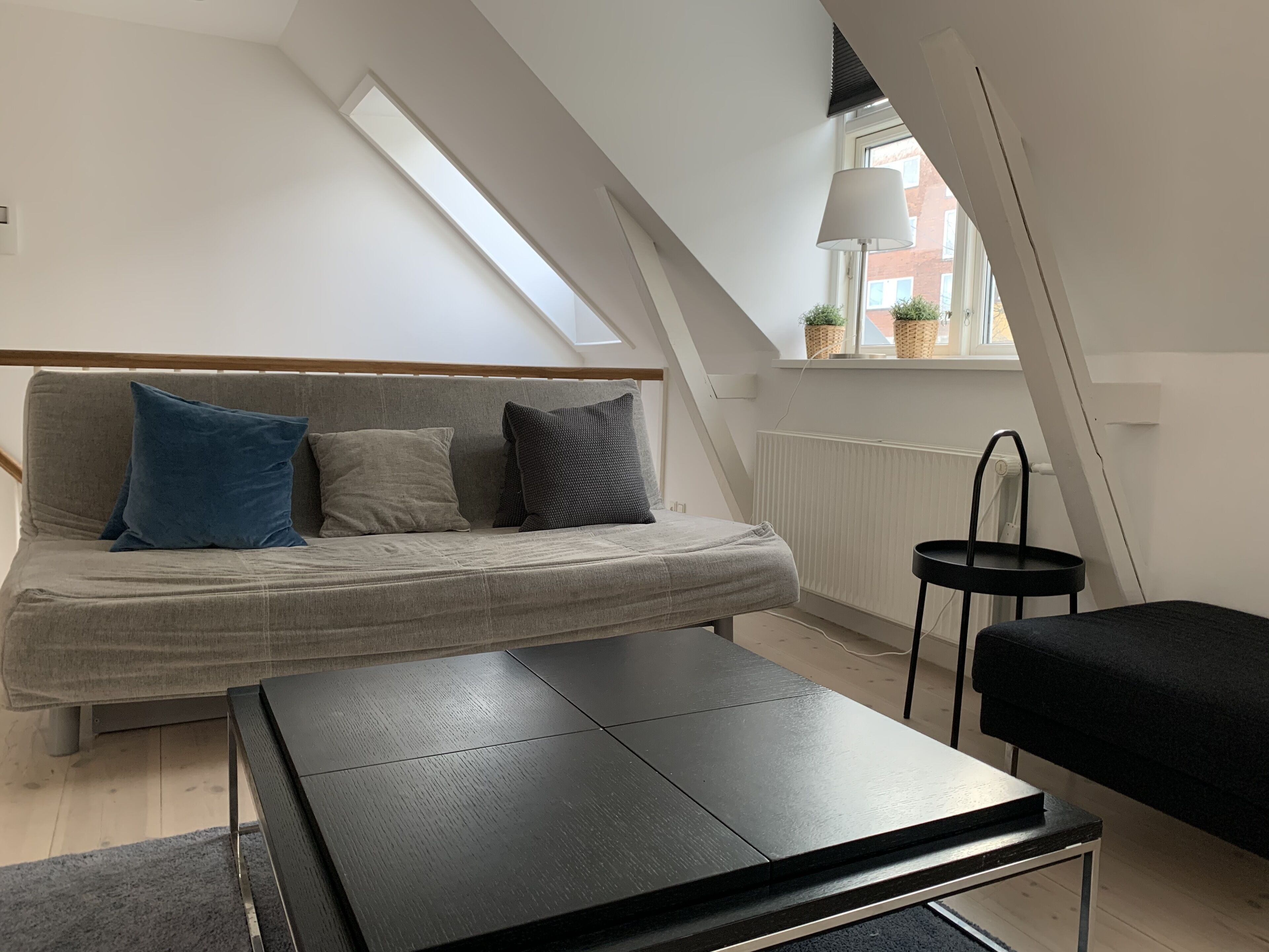 A loft-style bedroom with a sofa bed, a small table, and a window with a view of buildings.
