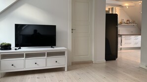 Moderni olohuone, jossa on valkoinen TV-taso ja siihen sijoitettu litteänäyttöinen televisio, ovi, joka johtaa keittiöön, sekä kalteva katto.