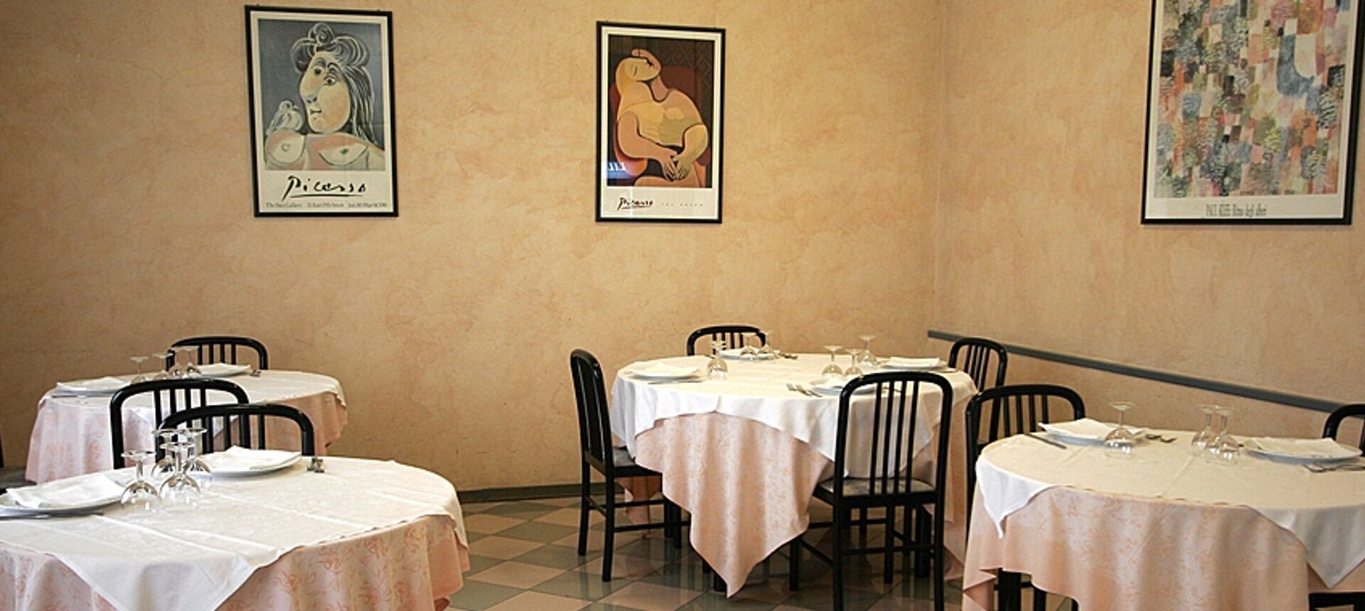 A dining area with tables set for two, chairs, and framed artwork on the walls.