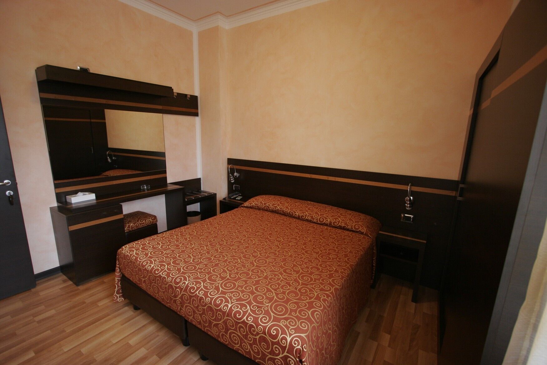 Une chambre d’hôtel de taille réduite, comprenant un lit, un bureau avec un miroir, un petit banc et une armoire.