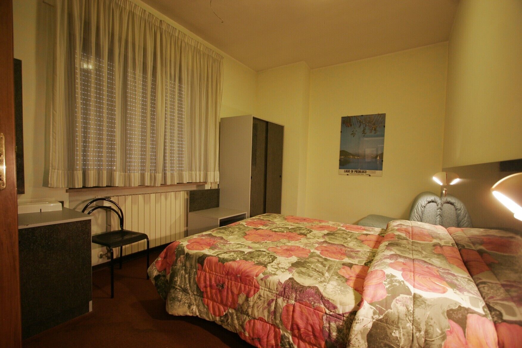 A hotel room with a bed, a desk, a chair, a radiator, and a poster on the wall.
