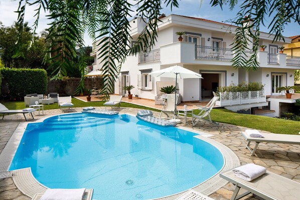A swimming pool with lounge chairs and umbrellas, surrounded by a well-maintained garden and a two-story building with balconies.