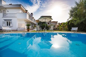 A swimming pool with lounge chairs and a sun umbrella, surrounded by a white building with balconies and potted plants.