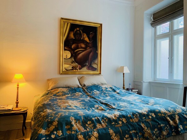 A bedroom with a large bed, a painting on the wall, a window with blinds, and a nightstand with a lamp. - Best Stay Copenhagen - Nyhavn 8 (Copenhagen)