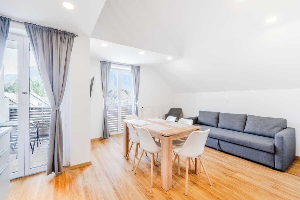 A modern living room with a wooden dining table, white chairs, a grey sofa, and a sliding glass door leading to a balcony.