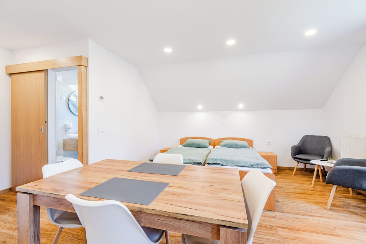 A modern, well-lit room with a wooden dining table, two beds with green bedding, a grey armchair, and a small white table.
