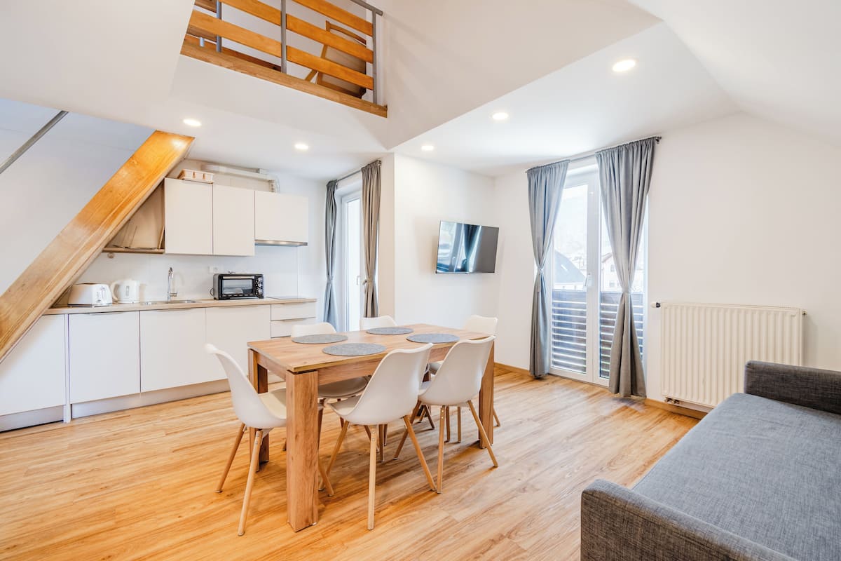 A modern kitchen with white cabinets, a wooden dining table with four chairs, a TV mounted on the wall, and a staircase with a wooden railing.